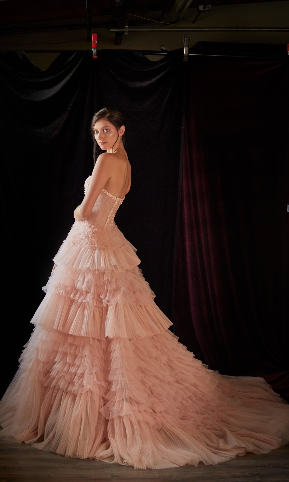 A woman wearing the Strapless Tiered Long Prom Ball Gown A1017, a light pink strapless ruffled tulle dress, stands in front of a dark curtain and looks over her shoulder.